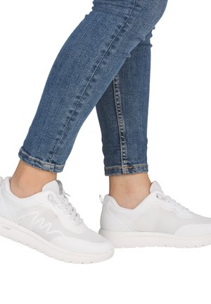 Person, die blaue Skinny-Jeans und weiße sportliche Sneakers trägt, steht mit einem leicht angehobenen Fuß.