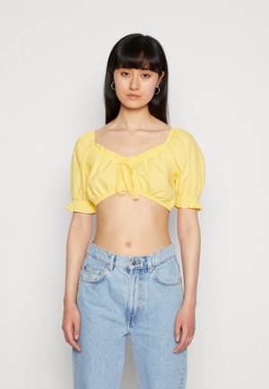 Blusa amarela cropped com mangas curtas bufantes, decote franzido e detalhe de laço na frente, combinada com jeans de cintura alta em azul claro.
