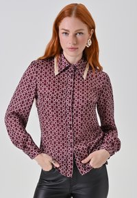 Camicia abbottonata color bordeaux con un motivo a catena bianca. Presenta un colletto classico e accenti di colletto color crema, maniche lunghe e un orlo svasato.