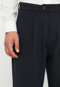 Námorně modré kalhoty s proužky, s knoflíkovým zapínáním a sklady vpředu. Vyrobeny z hladké tkaniny s upraveným střihem, zobrazeny s bílou košilí.