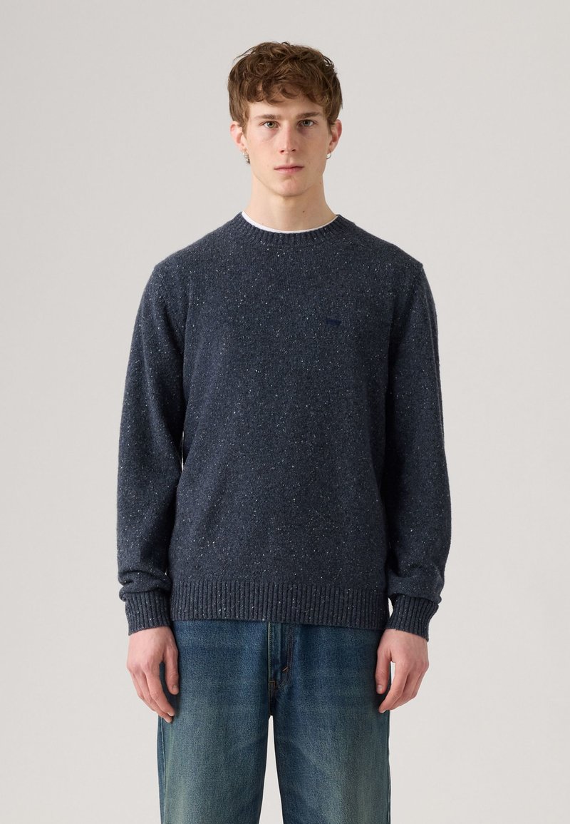 Mörkblå stickad tröja med en texturerad design, ribbade ärmslut och fåll, med ett subtilt spräckligt mönster. Bärs över ljusblå jeans.