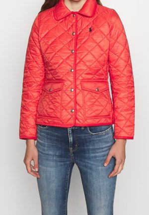 Vrouw draagt een rode gewatteerde jas met drukknopen en twee klepzakken aan de voorkant, gecombineerd met een blauwe spijkerbroek, staand tegen een effen achtergrond.