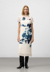 Vrouw in een crèmekleurige geplooide midi-jurk met blauwe abstracte patronen en zwarte leren kniehoogte laarzen, staand op een grijze vloer.
