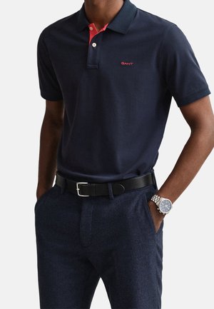 Homme portant un polo bleu marine avec des touches rouges, un pantalon sombre, une ceinture noire et une montre-bracelet argentée avec un cadran bleu, mains dans les poches.