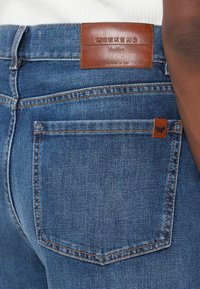Gros plan sur la poche arrière d'un jean en denim bleu avec une étiquette en cuir marron portant la mention « WEEKEND MaxMara » et un onglet avec logo papillon sur le bord de la poche.
