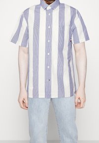 Koszula z krótkim rękawem z pionowymi niebiesko-białymi paskami, kołnierzykiem na guziki oraz małą kieszenią na piersi, wykonana z lekkiego materiału.