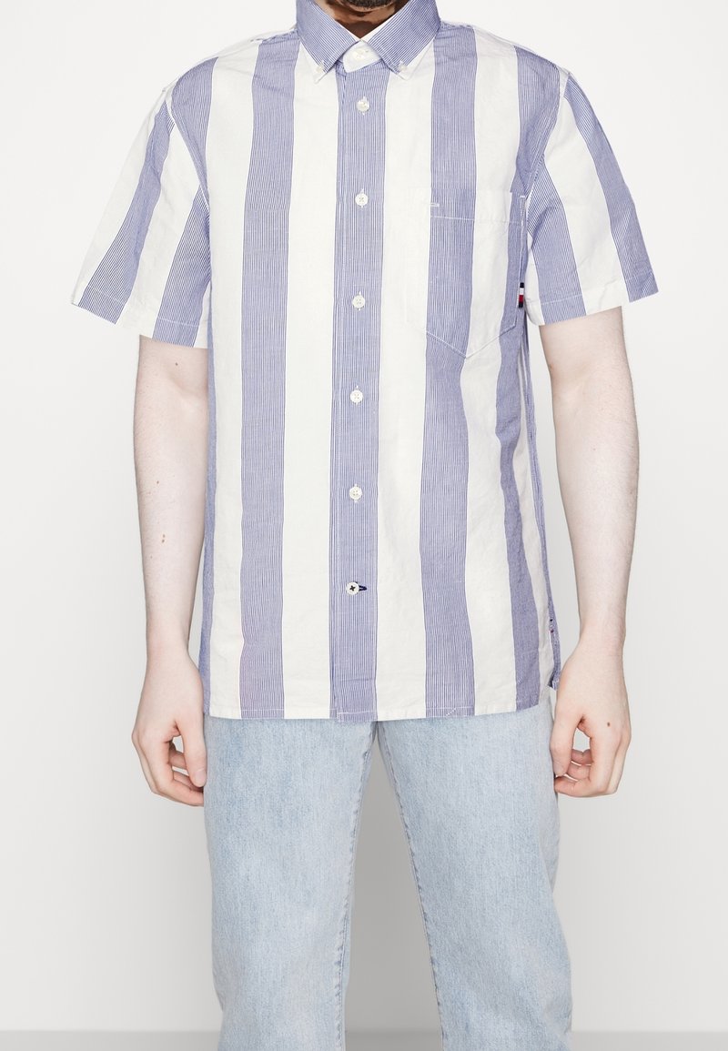 Koszula z krótkim rękawem z pionowymi niebiesko-białymi paskami, kołnierzykiem na guziki oraz małą kieszenią na piersi, wykonana z lekkiego materiału.