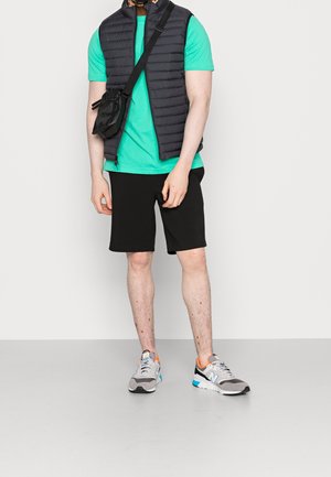 Man die een turquoise T-shirt, een zwart doorgestikt vest, zwarte korte broek, grijze sneakers en een zwarte crossbodytas draagt, staand tegen een witte achtergrond.