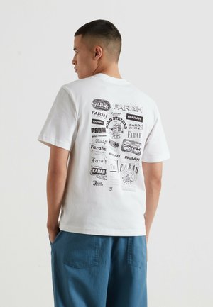 Mężczyzna o krótkich ciemnych włosach, ubrany w białą koszulkę z wieloma czarnymi logotypami Farah na plecach oraz niebieskie spodnie.