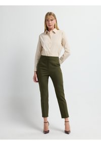 Beige overhem met een gestructureerde kraag, gecombineerd met olijfgroene op maat gemaakte broek en doorzichtige blokhak schoenen. Gladde texturen in het hele ensemble.