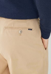 Beige broek van katoen, met een achterzak met knoopsluiting en een klein zwart "HACKETT" label. Subtiele textuur, nauwsluitende pasvorm.