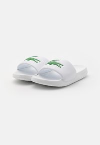 Sandales à glissière blanches en matériau synthétique avec un logo crocodile vert sur la sangle. Semelle texturée pour une bonne adhérence et un confort optimal.
