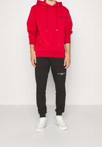 Sudadera de algodón roja con bolsillo frontal y capucha con cordón, combinada con pantalones de chándal negros que tienen texto en blanco. Usada con zapatillas blancas.