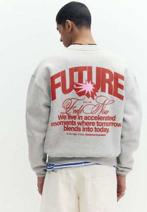 Mladi moški zadaj v sivem puloverju z velikim rdečim napisom „FUTURE“ in citatom o času na hrbtu.