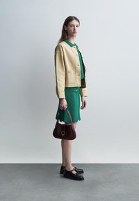 Vrouw in een groen gestructureerde jurk met een beige jas, ze draagt een donkerrode handtas en zwarte schoenen, staande tegen een effen witte muur.