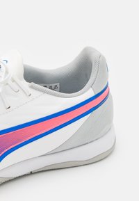 Zapatilla deportiva blanca con detalles grises, franja lateral en rosa y azul, talón de malla texturizada y diseño de puntera redondeada. Cordones y amortiguación visibles.