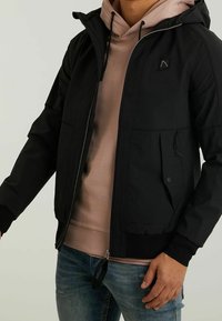 Homme portant une veste noire zippée par-dessus un sweat à capuche beige et un jean bleu, debout avec une main détendue sur le côt é.