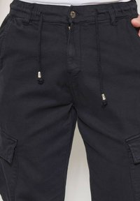 Pantalons cargo noirs avec une taille à cordon de serrage, fermeture à bouton, poches latérales et tissu texturé. Présente des détails de couture subtils.