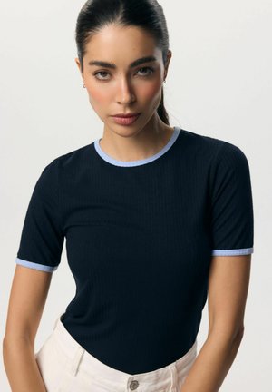 Kobieta z ciemnymi włosami związanymi do tyłu, ubrana w dopasowany granatowy, krótki rękaw z jasnoniebieskim obszyciem i białe spodnie na jednolitym tle.