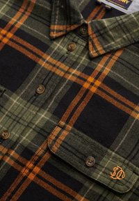 Green and black plaid flannel shirt with orange accents, featuring a button-down collar, chest pocket, and logo embroidery on the pocket.