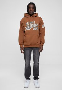 Felpa marrone con tasca frontale, caratterizzata da un design grafico e il numero 05. Abbinata a jeans grigi scuri e sneaker bianche.