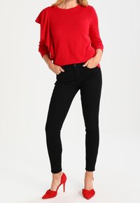 Femme portant un haut rouge à manches longues avec détail à volants, jean skinny noir et chaussures à talons rouges, debout les mains dans les poches.