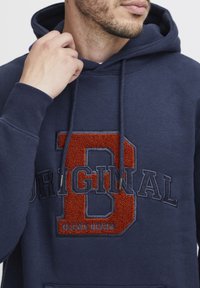 Navy kék pamut kapucnis pulóver, amelyen egy nagy, texturált narancssárga "D" és a "ORIGINAL" szó sötétkék hímzéssel található. Állítható húzózsinór a kapucnin.