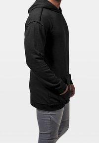 Sudadera con capucha negra de acabado texturizado, con bolsillos laterales, puños acanalados y un corte relajado. Llevada sobre pantalones gris claro.