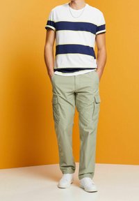 Man in witte sneakers, lichtgroene cargobroek en een wit T-shirt met navy en gele horizontale strepen tegen een oranje achtergrond.