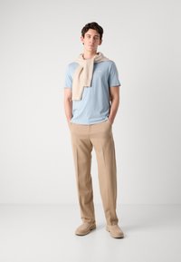 Maglietta blu chiaro con collo a giro e logo, maglione beige appoggiato sulle spalle, pantaloni beige su misura e scarpe di tonalità chiara. Texture di tessuti lisci.