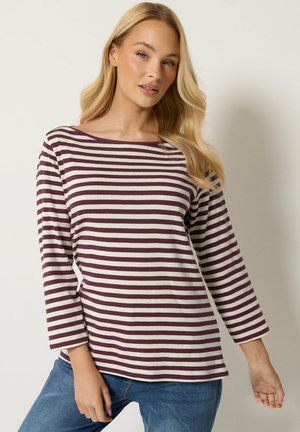 Donna bionda che indossa una maglia a righe bianche e rosso scuro a maniche lunghe e jeans blu, in piedi davanti a uno sfondo chiaro e uniforme.