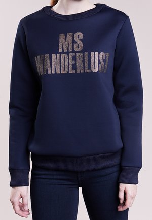 Granatowa bluza z błyszczącym srebrnym napisem „MS WANDERLUST”, noszona przez osobę w ciemnych dżinsach, stojącą na jednolitym tle.