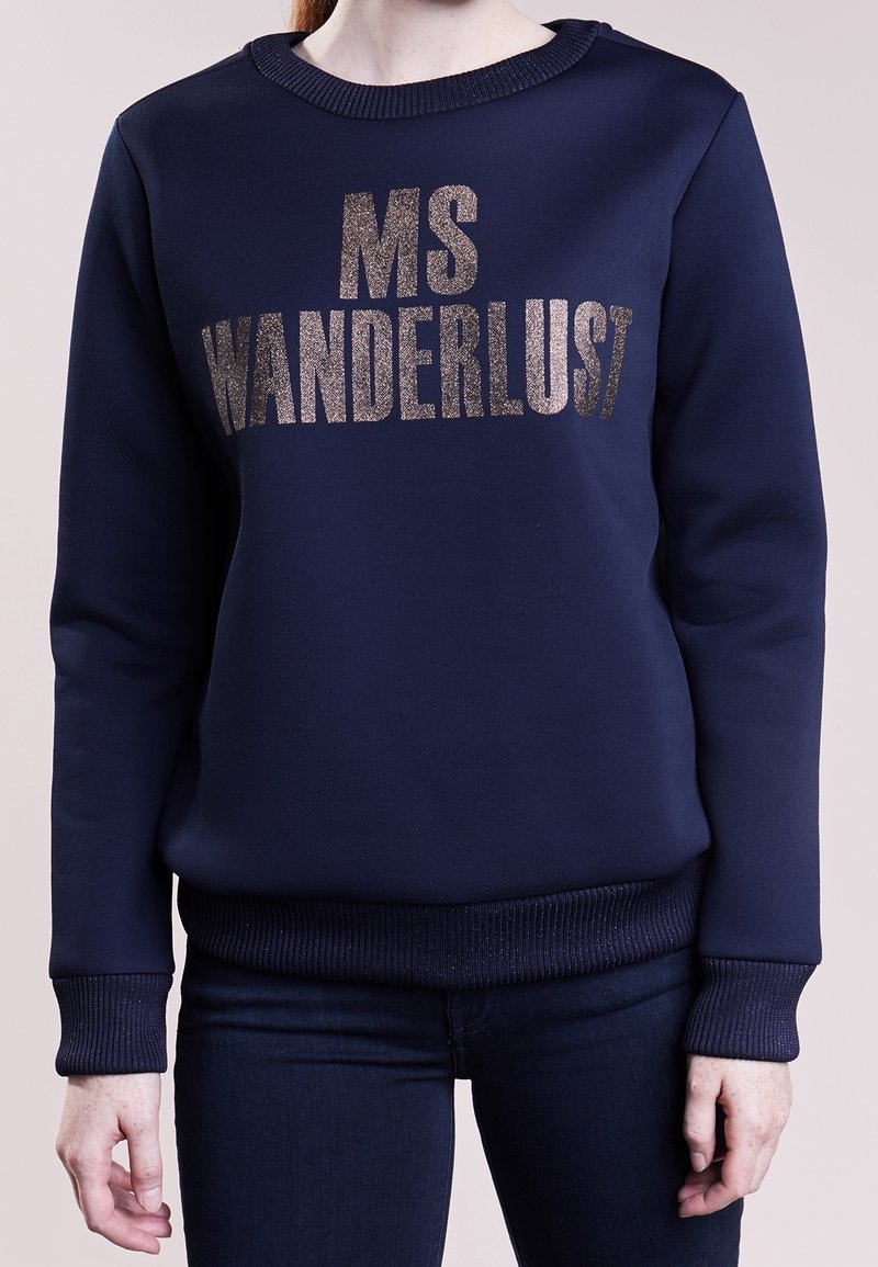 Sweat-shirt bleu marine avec texte argenté scintillant "MS WANDERLUST" porté par une personne en jean foncé, debout devant un arrière-plan uni.