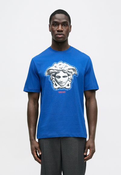 Modré bavlnené tričko s veľkým grafickým zobrazením Medusinovej hlavy v strede, ohraničené vzorom gréckeho kľúča, s textom „VERSACE“ pod tým.
