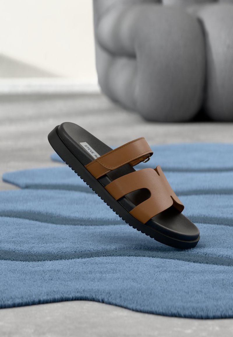 Sandale mule en cuir marron avec deux larges brides, semelle noire, semelle intérieure rembourrée et semelle extérieure en caoutchouc texturé, posée sur un tapis bleu.