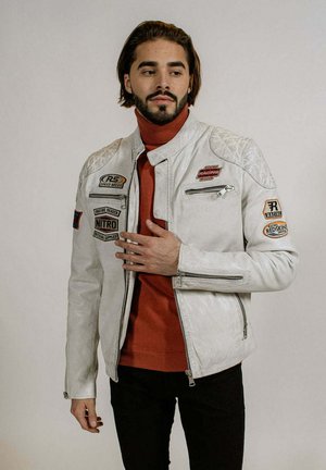 Veste de moto en cuir blanc avec des accents d'épaules matelassés, plusieurs patchs brodés et des poches zippées, portée par-dessus un col roulé rouge.