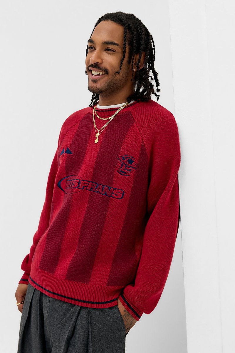 Camisola vermelha de malha com riscas verticais, gola e punhos canelados, com um logótipo e texto em azul-marinho. Combinada com calças cinzentas plissadas.