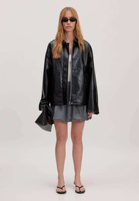 Zwarte leren oversized jas met een subtiele glans, gecombineerd met een grijze gerimpelde minirok. Versterkt met een zwarte clutch en platte sandalen.