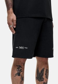 Zwarte sweatshorts van textiel met een textuur, met witte geborduurde tekst "British No1 Design" op het linkerbeen.