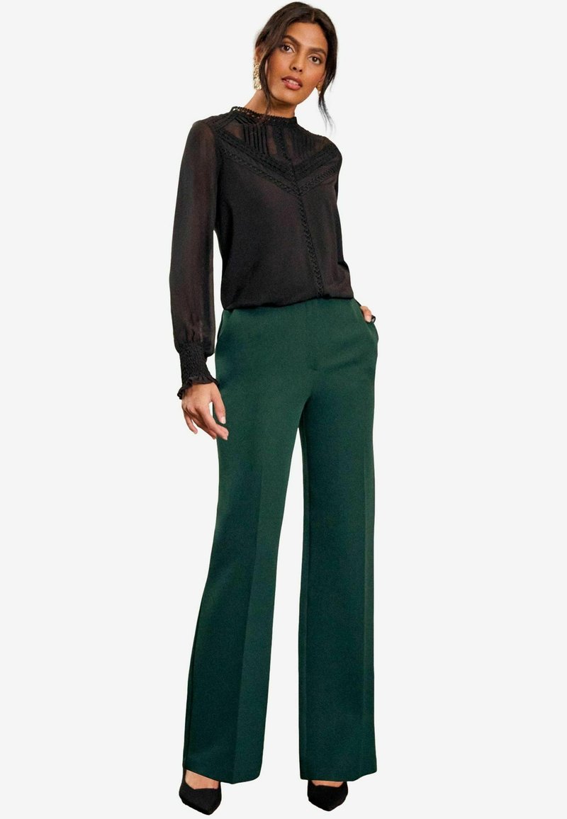 Schwarze durchsichtige Bluse mit Spitzenverzierungen, kombiniert mit grünen, hoch taillierten, weiten Hosen und schwarzen High-Heels.