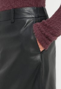 Χέρι στη τσέπη της μαύρης δερμάτινης φούστας που φοριέται με φουντωτό μακρυμάνικο μπορντό πουλόβερ, εμφανίζεται από τη μέση μέχρι το μέσο του μηρού.