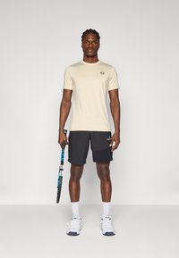 Μπεζ αθλητικό μπλουζάκι με κοντά μανίκια και στρογγυλή λαιμόκοψη, συνδυασμένο με μαύρες βερμούδες με πλαϊνές τσέπες, κρατώντας μια ρακέτα τένις.