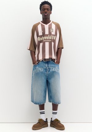 Nuori mies, joka käyttää ruskeaa ja vaaleanpunaista raidallista urheilupaitaa, pitkät farkkushortsit, joissa näkyvä teksti, valkoiset sukat mustilla raidoilla ja ruskeat kengät.