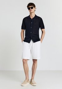Mann in einem schwarzen kurzärmligen Hemd mit Knöpfen, weißen knielangen Shorts, beigen Slippern und schwarzen Sonnenbrille, der mit den Händen in den Taschen steht.
