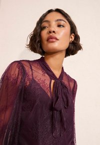 Žena s krátkymi vlnitými vlasmi, oblečená v priesvitnej fialovej čipkovanej blúzke s golierom na mašľu, ktorá sa pozerá mierne nahor proti jednoduchému pozadiu.