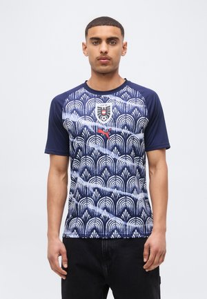 Νέος άνδρας φορώντας μπλε-σκούρα και λευκή ποδοσφαιρική φανέλα με κόκκινο λογότυπο Puma και μαύρο παντελόνι, στέκεται μπροστά σε απλό φόντο.