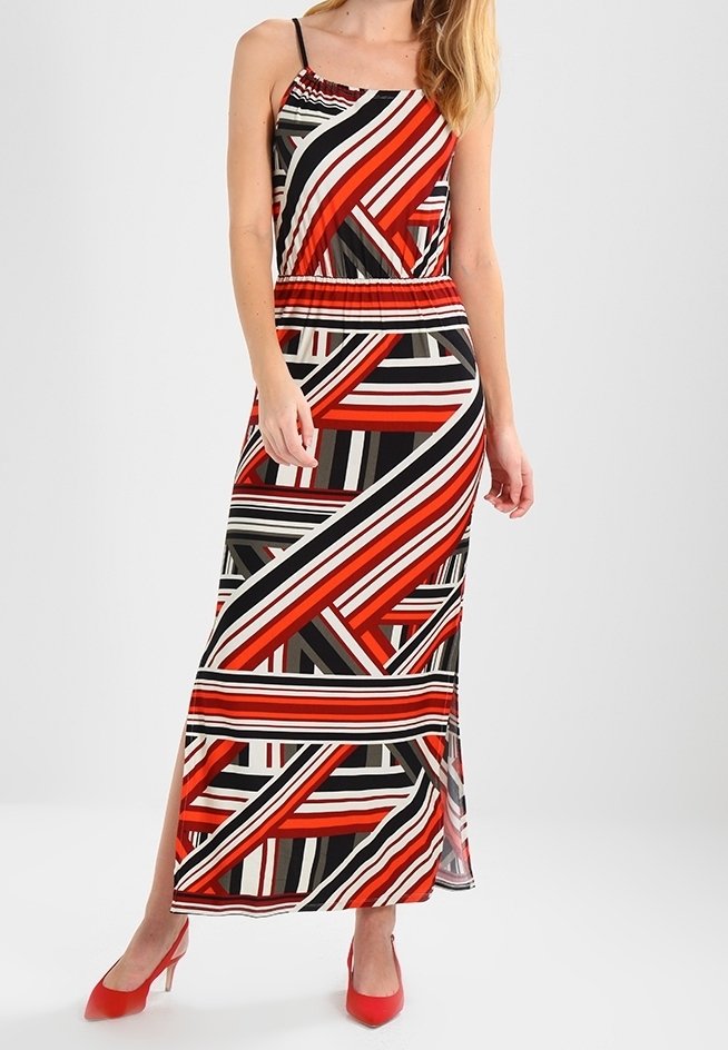 Kvinna klädd i en ärmlös klänning som når anklarna, med geometriska ränder i röd, svart, vit och beige, kombinerat med röda klackar.