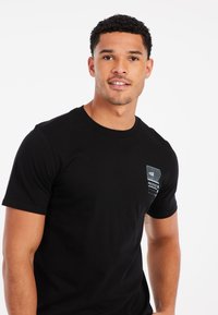 Schwarzes Baumwoll-T-Shirt mit Rundhalsausschnitt und kurzen Ärmeln, das einen kleinen grafischen Druck in Blau und Weiß auf der linken Brustseite aufweist.
