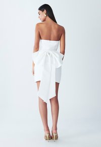 White strapless dress featuring a large bow at the back, short length, smooth fabric, and a straight silhouette. High-heeled shoes are worn.