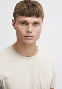 Beige crew-neck t-shirt med en blød tekstur. Modellen har kort brunt hår og blå øjne, med minimal synlig tilbehør.